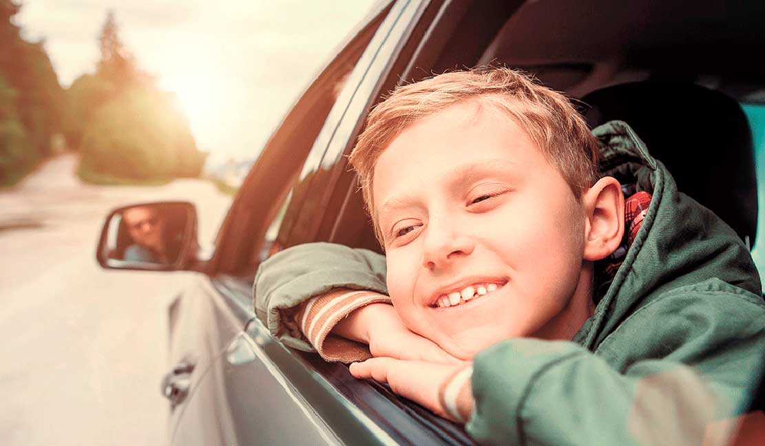 Hymyilevä poika nojaa autonsa ikkunaan ja katsoo ulos maisemia. Taustalla näkyy aurinkoinen tie ja auton sivupeilissä heijastuu kuljettaja.
