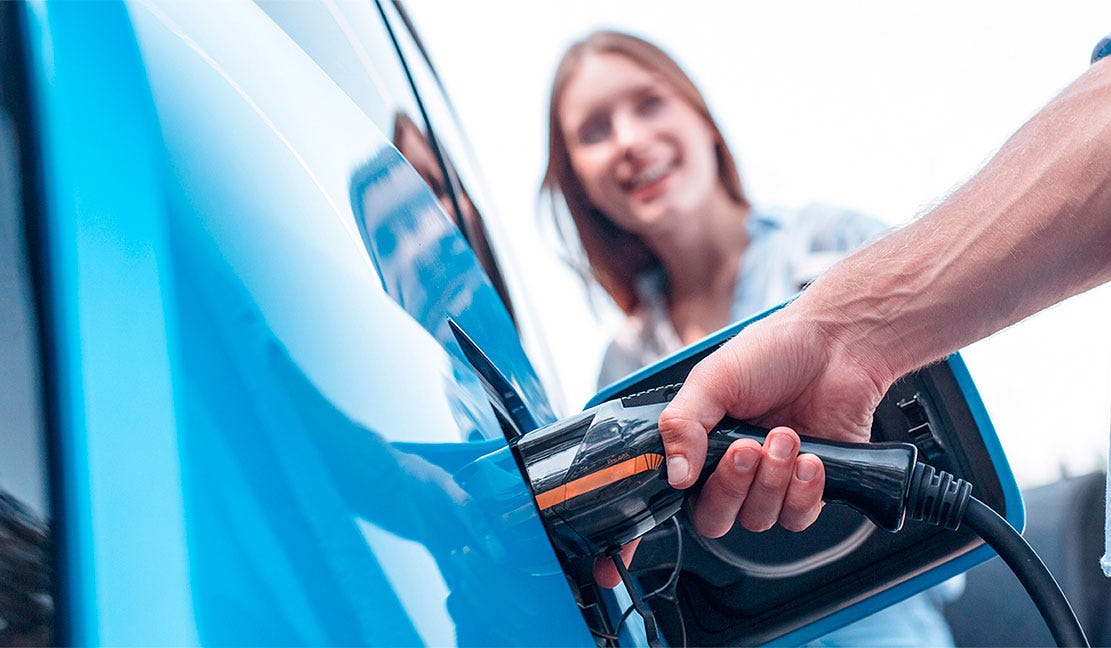 Lähikuva sähköauton lataamisesta. Käsi liittää latauskaapelin sinisen auton latausporttiin, taustalla hymyilevä nainen.