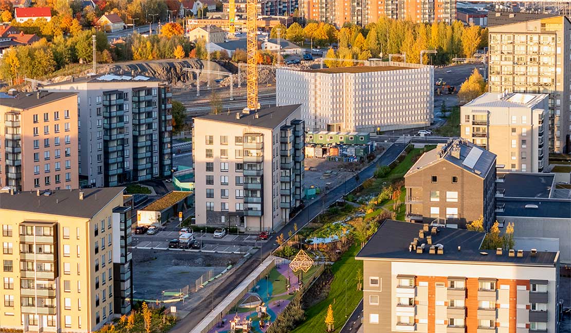 Vuokrattavat autopaikat Ilmakuva Kirstinpuiston kaupunkialueesta, jossa on useita kerrostaloja, vehreitä puistoalueita ja leikkipaikka. Taustalla näkyy Kirstinpuiston pysäköintitalo, josta on mahdollista vuokrata autopaikkoja.