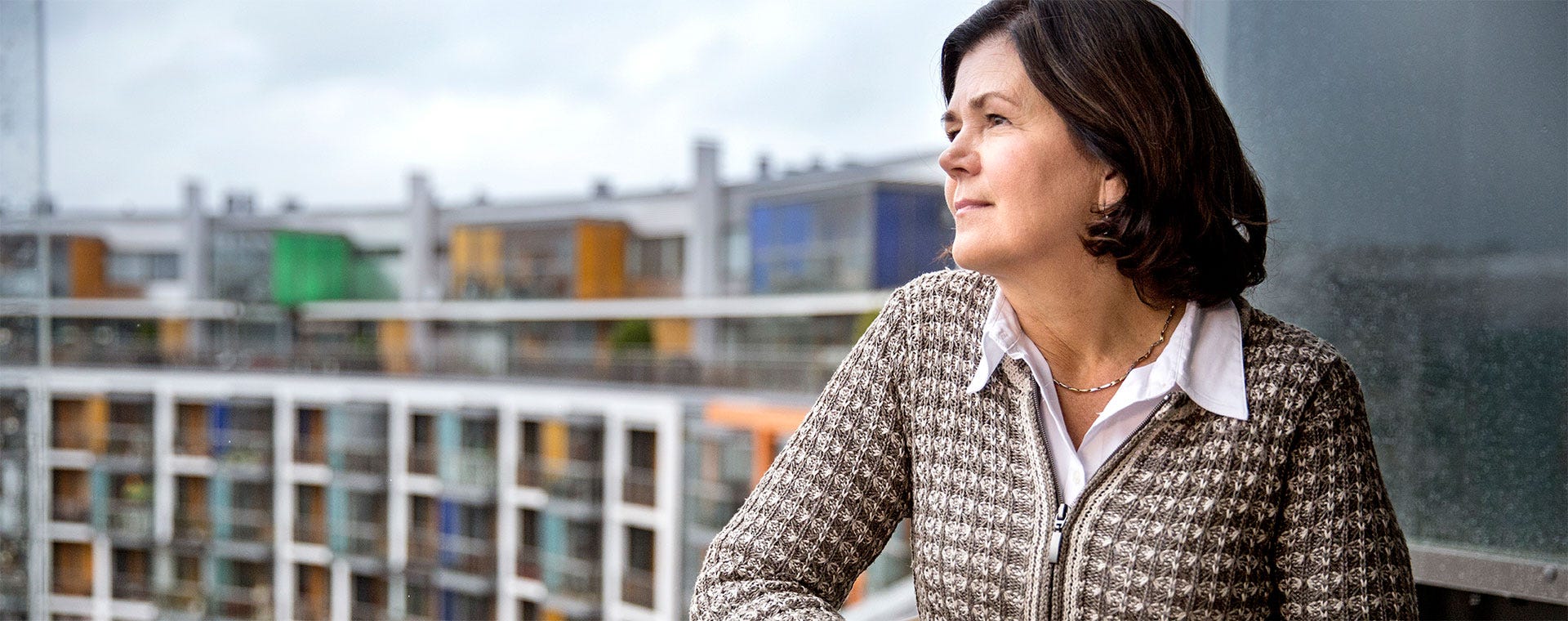 Nainen katselee rauhallisesti kaupunkimaisemaa parvekkeeltaan. Taustalla näkyy moderni kerrostalo, jossa on lasitetut parvekkeet ja värikkäitä yksityiskohtia. 
