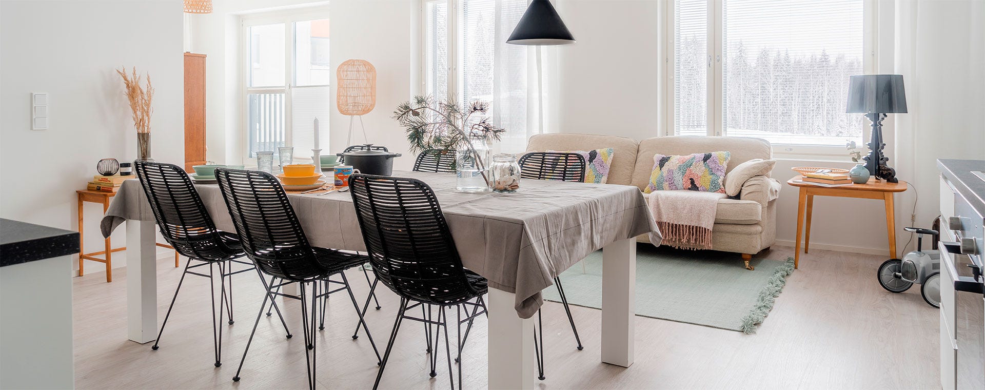 Valoisa ruokailu- ja oleskelutila, jossa pitkä ruokapöytä mustine tuoleineen, rento vaalea sohva värikkäin tyynyin sekä suurista ikkunoista avautuva talvinen metsämaisema.