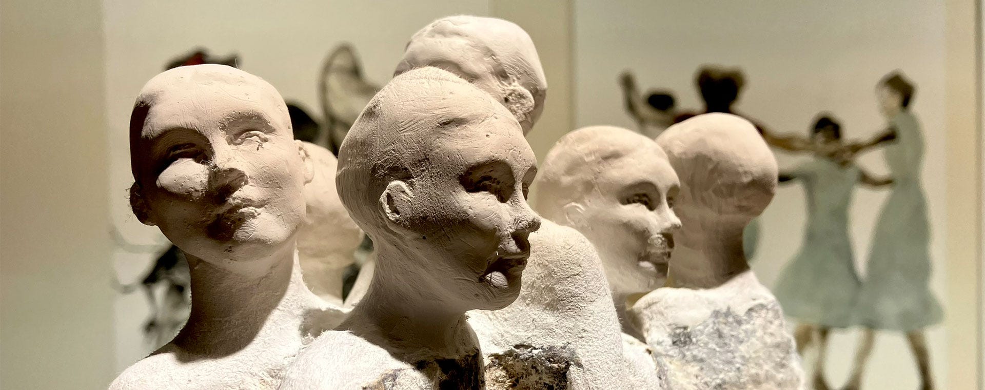 Lähikuvassa joukko vaaleasävyisiä saviveistoksia, jotka esittävät ihmisten päitä ja hartioita. Taustalla näkyy utuinen maalaus tanssivista hahmoista, mikä luo kuvallisen yhteyden liikkeen ja hiljaisuuden välille.