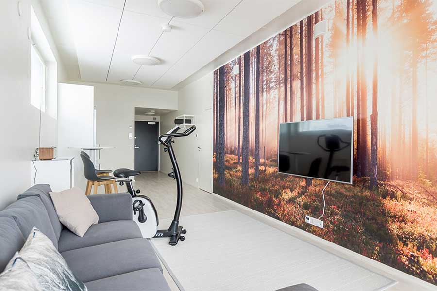 Kerhotila, jossa on sohva, kuntopyörä, televisio ja metsää esittävä seinäkuva.