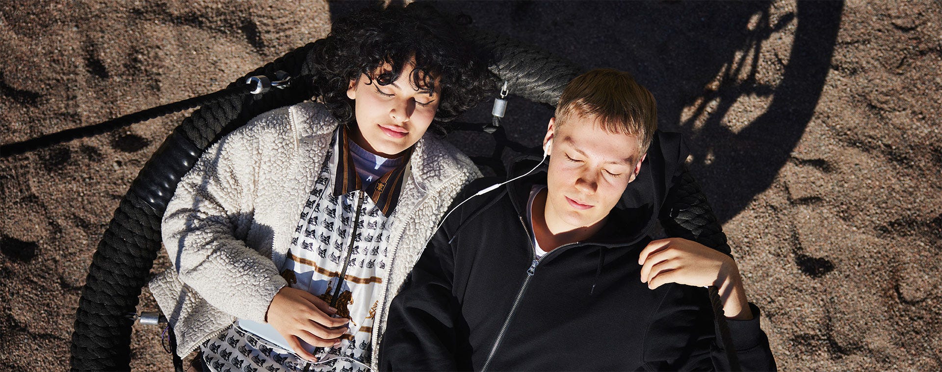 Kaksi nuorta lepää suuren keinun päällä silmät suljettuina auringonpaisteessa. Heidän ympärillään hiekkainen leikkipaikka, ja varjot piirtävät kuvioita maahan. Toinen kuuntelee musiikkia kuulokkeilla, ja molemmilla on rento ja rauhallinen ilme.