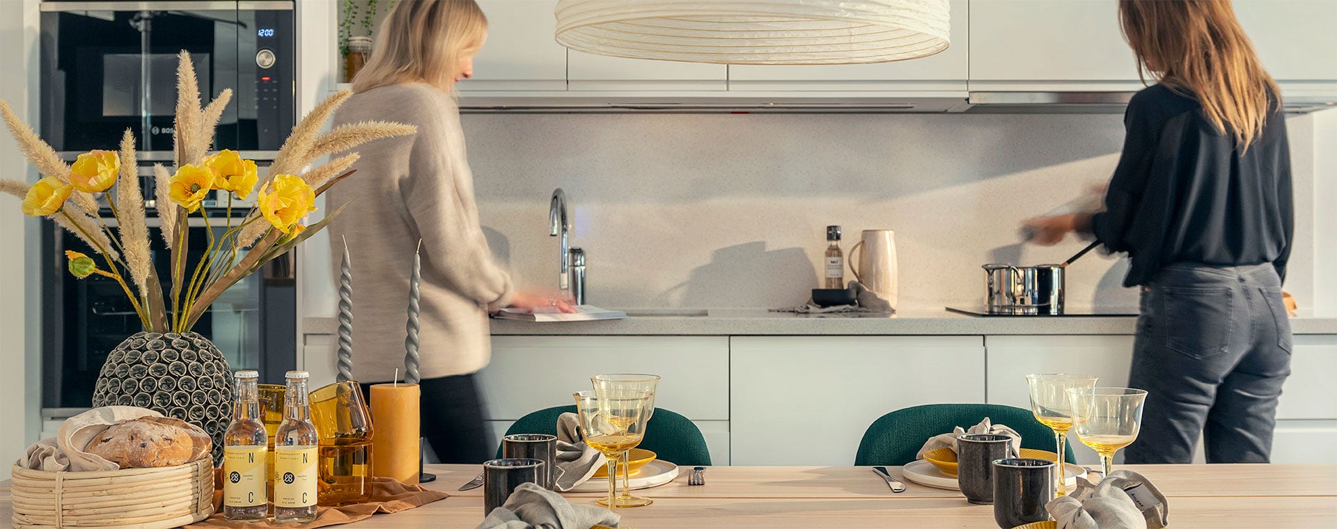 Kaksi naista valmistautuu ateriaan modernissa keittiössä. Toinen pesee käsiään tiskialtaan äärellä, kun taas toinen sekoittaa ruokaa kattilassa hellalla. Etualalla on kauniisti katettu ruokapöytä, jossa on keltaisia yksityiskohtia, viinilasit, kangasservetit ja koristeellisia kynttilöitä.