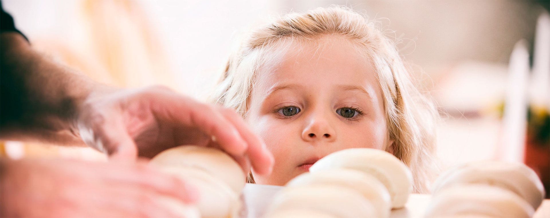 Pieni lapsi katsoo uteliaana leipomisvaiheessa olevia pullia. Etualalla aikuisen kädet asettelevat taikinapalloja leivinpaperille. Pehmeä valo tuo lämpöä keittiöhetkeen.