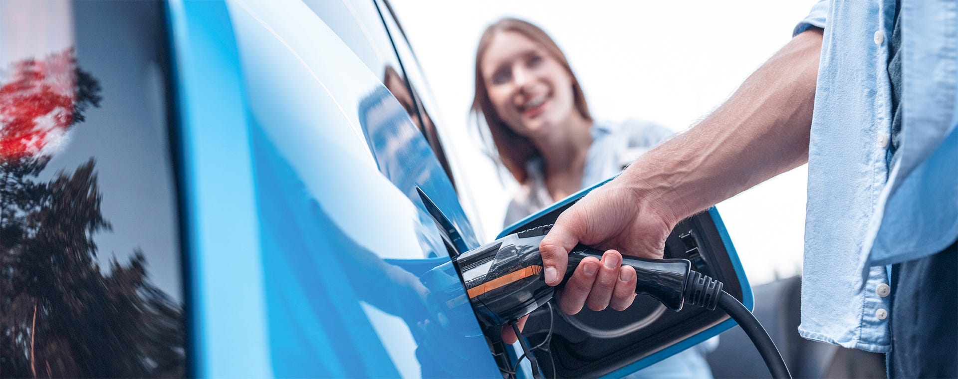 Lähikuva sähköauton lataamisesta. Käsi liittää latauskaapelin sinisen auton latausporttiin, taustalla hymyilevä nainen.
