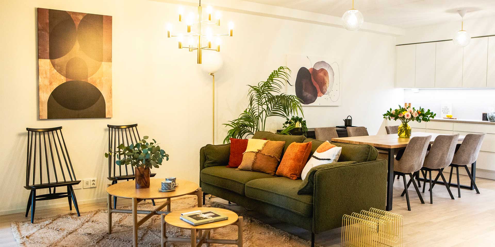 Lämminhenkinen ja tyylikäs olohuone, jossa vihreä sohva koristetyynyineen, kaksi pyöreää puista sohvapöytää sekä pehmeä matto. Seinillä abstraktia taidetta ja taustalla näkyy ruokailutila, jossa suuri puupöytä kukka-asetelmalla sekä verhoillut tuolit. Huoneessa on vehreitä viherkasveja ja tunnelmallinen valaistus.