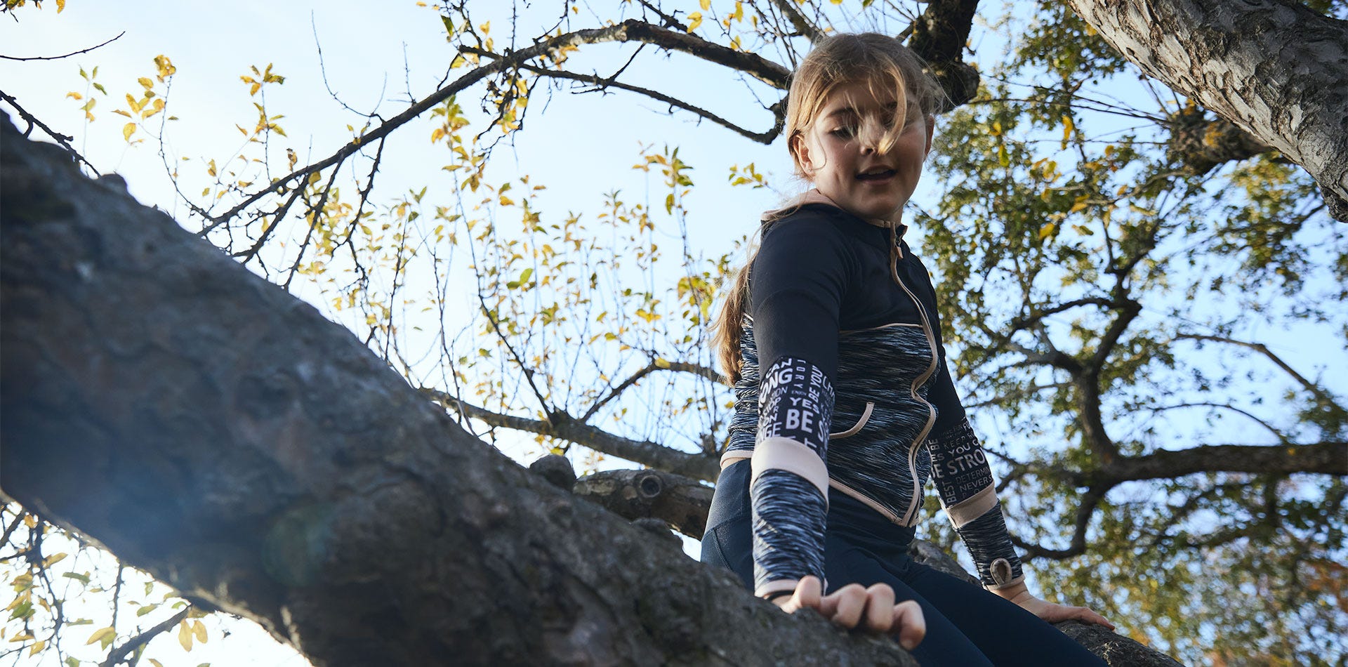 Lapsi kiipeämässä puussa syksyisessä maisemassa. Auringonvalo siivilöityy puun oksien läpi, ja taustalla näkyy keltaisia ja vihreitä lehtiä. Lapsi katsoo alas keskittyneesti ja pitää kiinni puunrungosta.