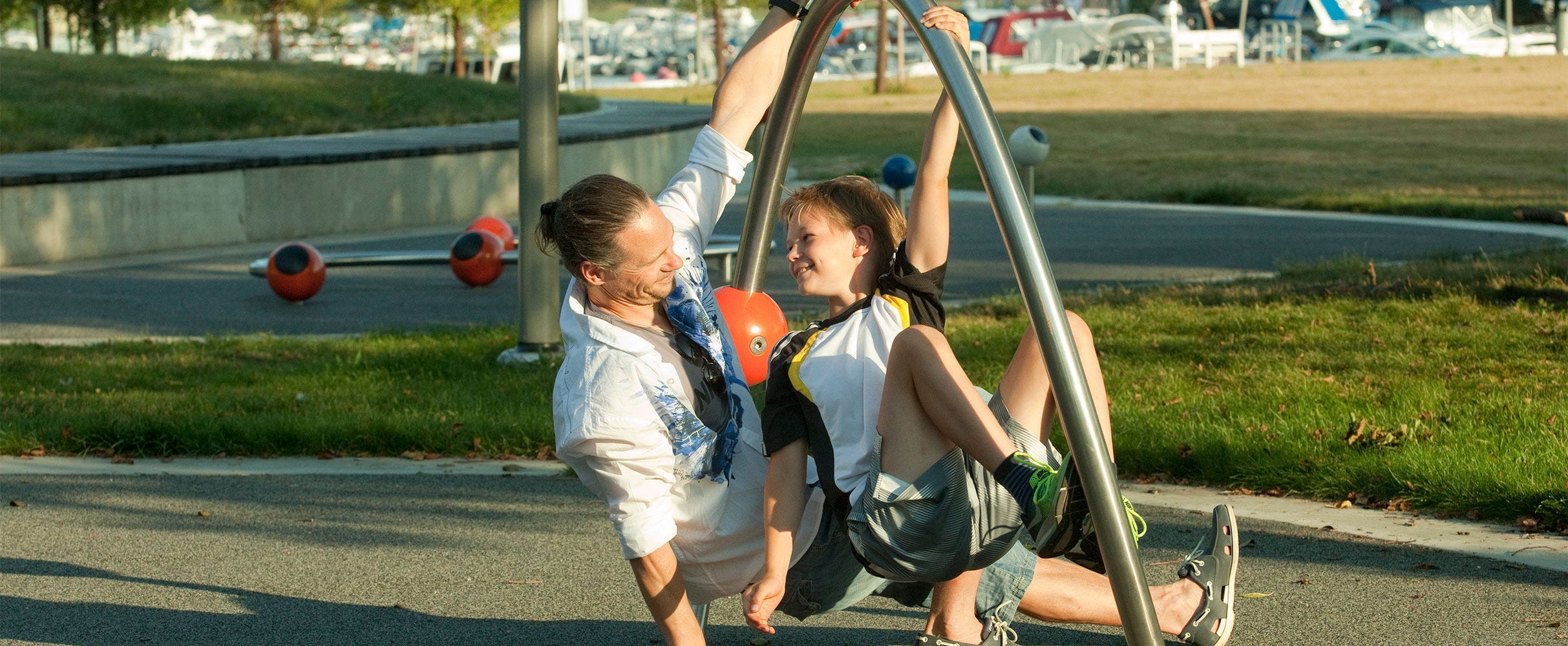 Isä ja poika leikkivät modernilla kiipeilytelineellä vehreässä puistossa. He nauravat yhdessä ja näyttävät nauttivan iloisesta ulkoiluhetkestä. Taustalla näkyy Otsolahden venesatama ja nurmialue aurinkoisena päivänä.