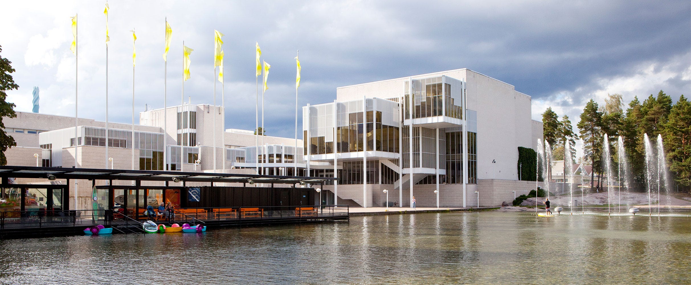 Espoon kulttuurikeskus on moderni valkoinen rakennus, jossa on paljon lasia ja korkeat ikkunat. Rakennuksen edustalla lipputankoja, pieni laituri ja kirkasvärisiä kajakkeja vedessä.