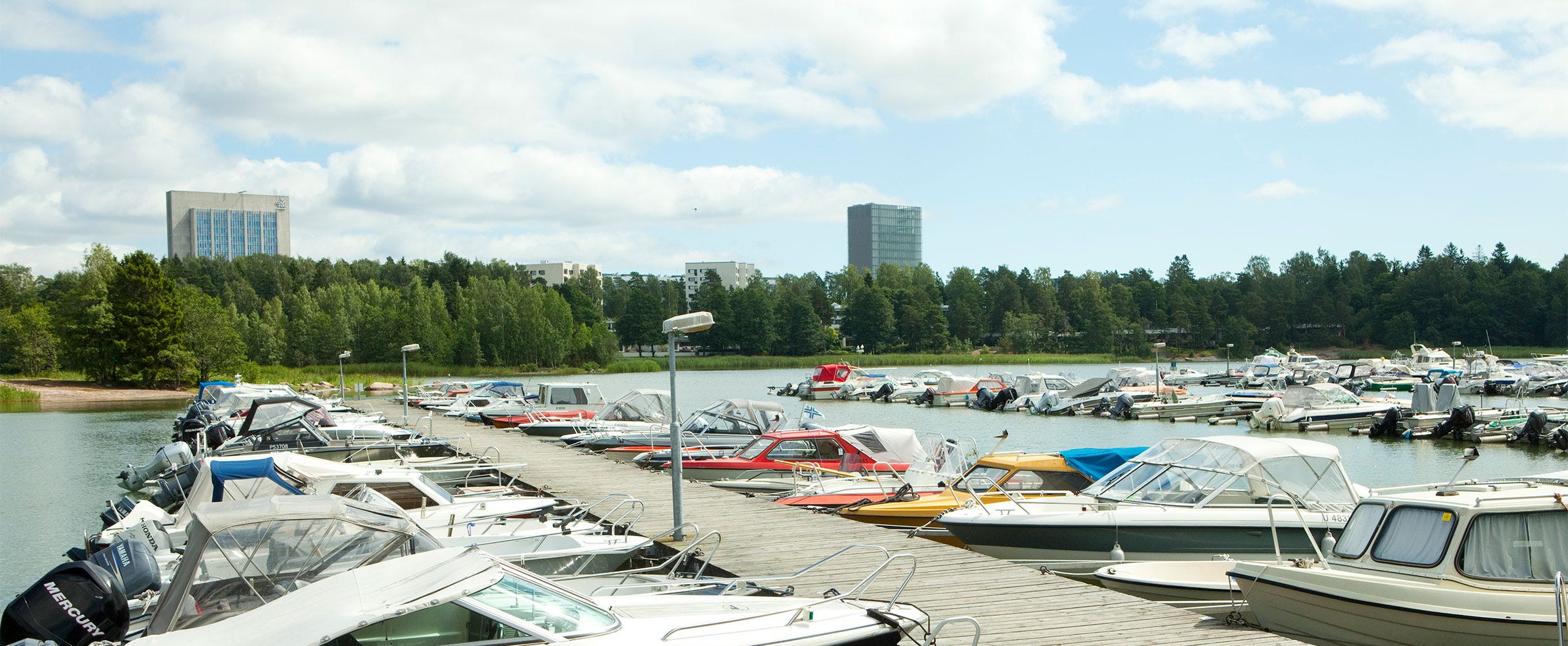 Otsolahden venesataman laituri, jonka molemmin puolin on pysäköityjä veneitä. Taustalla näkyy vehreää metsää, kerrostaloja ja kaksi korkeampaa rakennusta.
