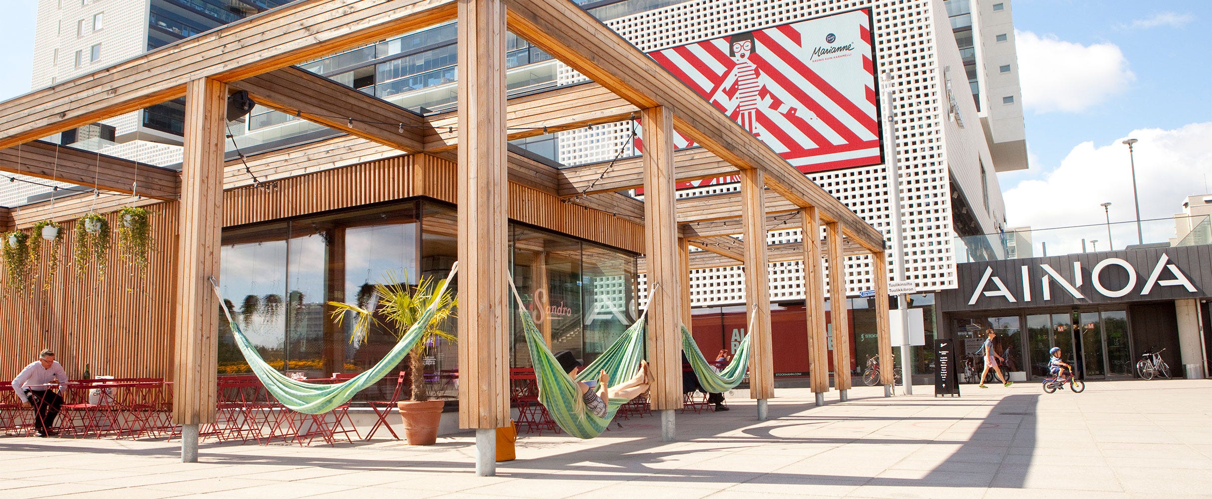 Kuvassa on AINOAn ostoskeskuksen ulkoterassi Espoossa. Puisen pergolarakenteen alla on värikkäitä riippumattoja ja punaisia pöytäryhmiä. 