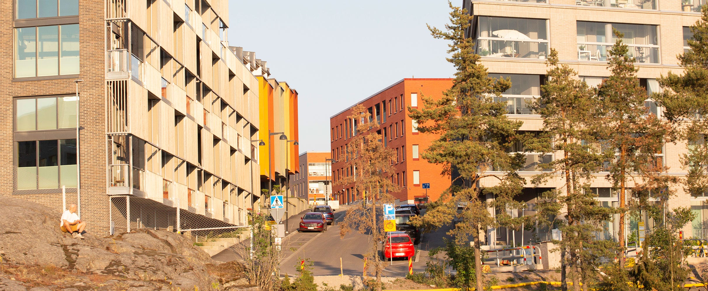 Katu Kruunuvuorenrannassa, jota reunustavat modernit kerrostalot. Rakennukset ovat värikkäitä ja oikealla lasiset parvekkeet erottuvat selkeästi. Kadun päässä näkyy punatiilinen rakennus. Metsäinen ympäristö kehystää kaupunkimaisemaa.