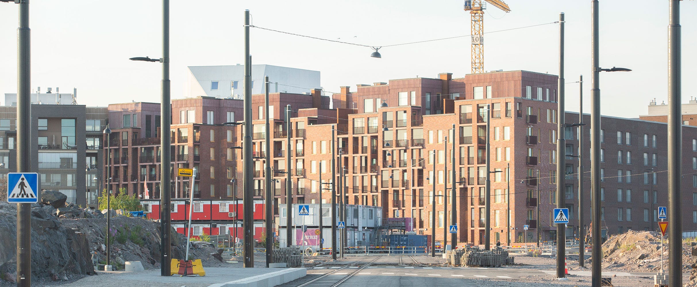 Kruunuvuorenrannassa punatiiliset kerrostalot nousevat korkeuksiin. Kadulla näkyy raitiovaunukiskot ja työmaa-aitoja. Rakennusnosturi kohoaa taustalla, ja valopylväät rajaavat katunäkymää.