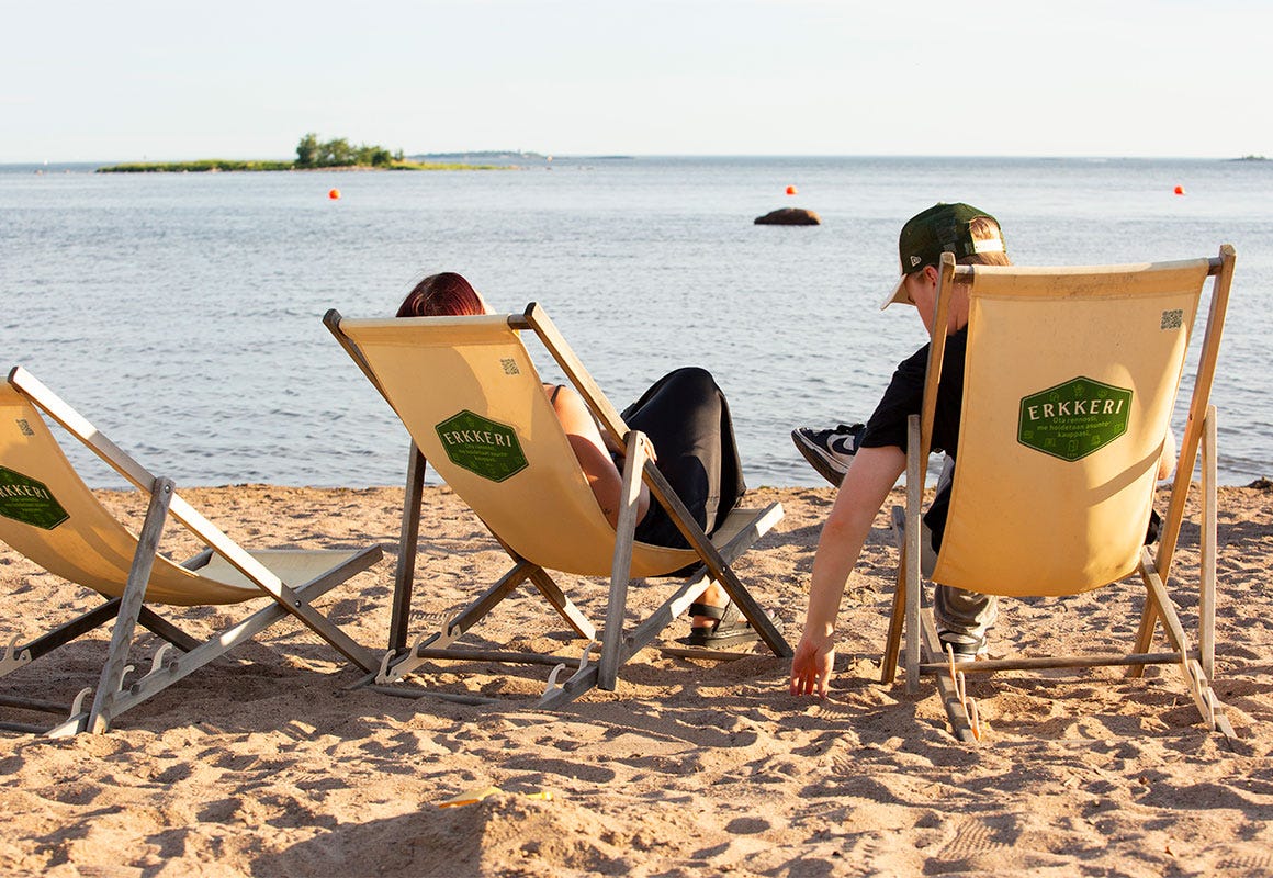 Kaksi ihmist&auml; istuu rantatuoleissa Lauttasaaren hiekkarannalla. Edess&auml; avautuu meri ja pieni saari.