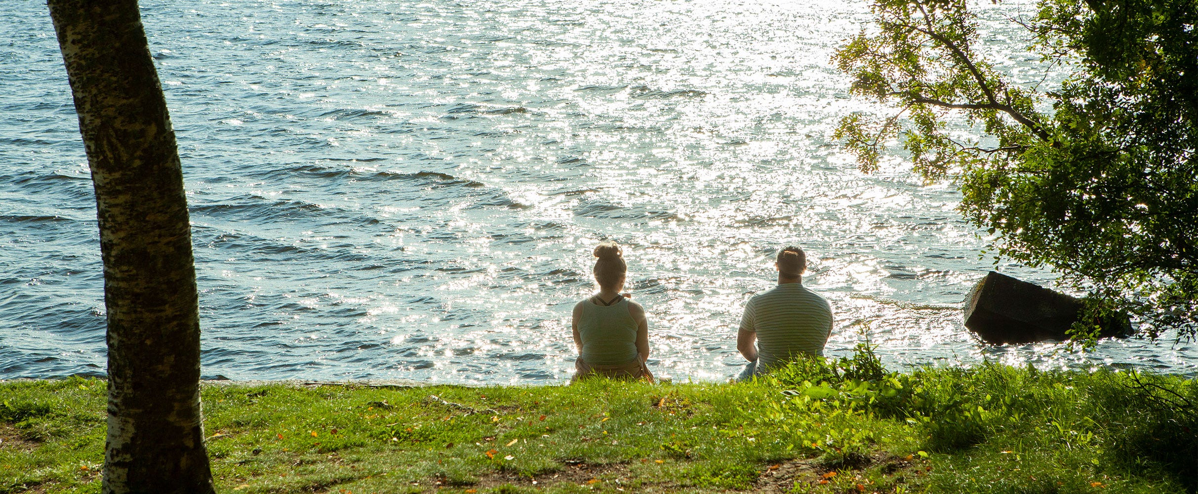 Kaksi henkilöä istuu nurmikolla Pyhäjärven rannalla nauttien auringonpaisteesta. Veden pinta kimaltelee kirkkaasti auringonvalossa ja ympärillä kasvaa vihreitä puita.