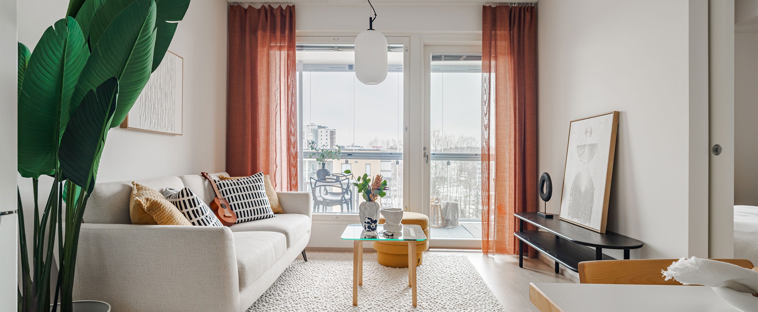 Valoisa olohuone, jossa on moderni sisustus. Valkoinen sohva koristetyynyineen, matala musta taso ja lasipöytä, jolla on kukkakimppu. Iso viherkasvi tuo vehreyttä. Ikkunasta avautuu näkymä lasitetulle parvekkeelle ja talvimaisemaan.