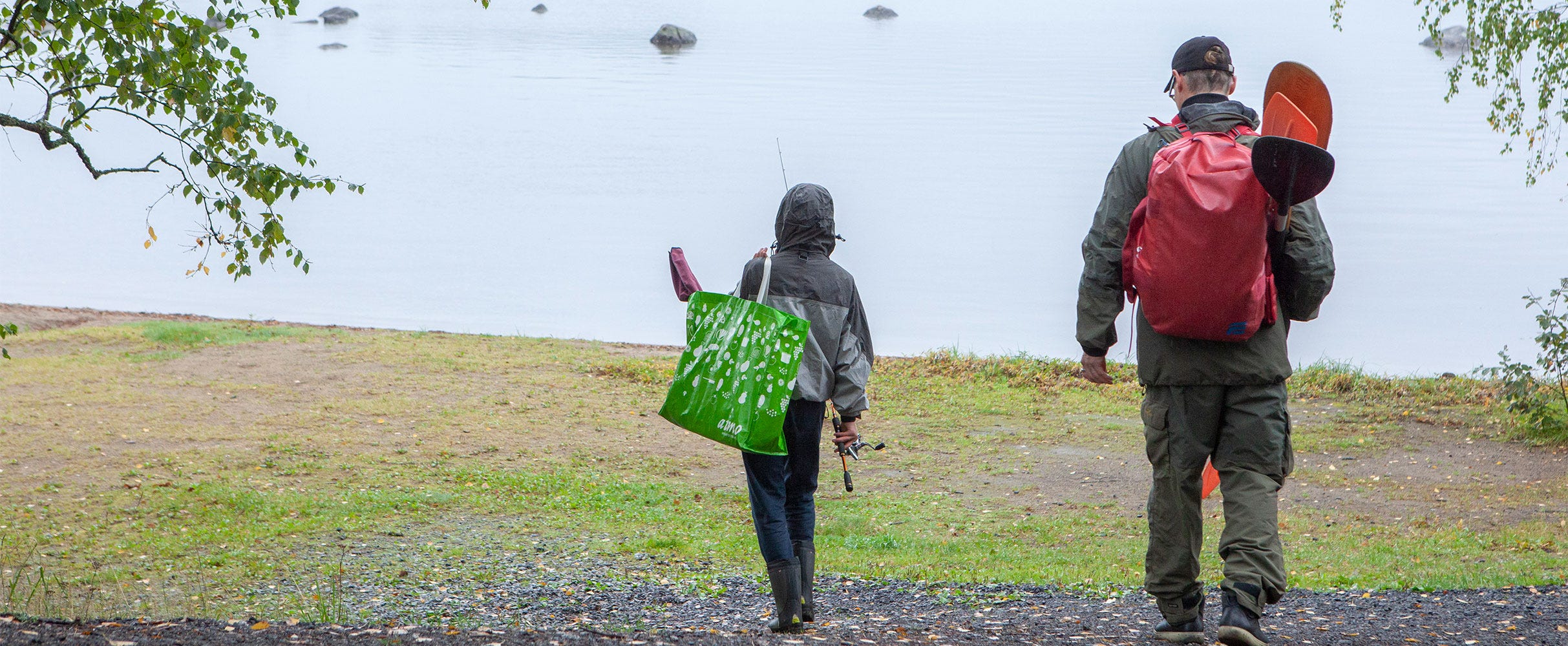 Kaksi henkilöä kävelee kohti järven rantaa sateisessa säässä. Toinen kantaa vihreää kassia ja kalastusvälineitä, toinen reppua ja melaa. 