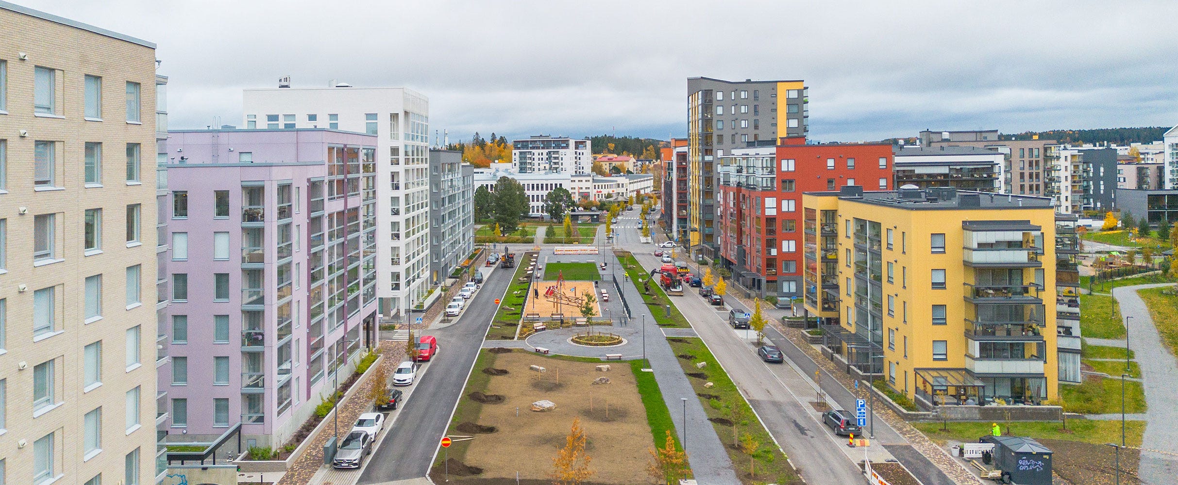 Ilmakuva modernista asuinalueesta, jossa värikkäät kerrostalot reunustavat leveää katua. Keskellä viheralue, leikkipuisto ja kävelyreittejä.