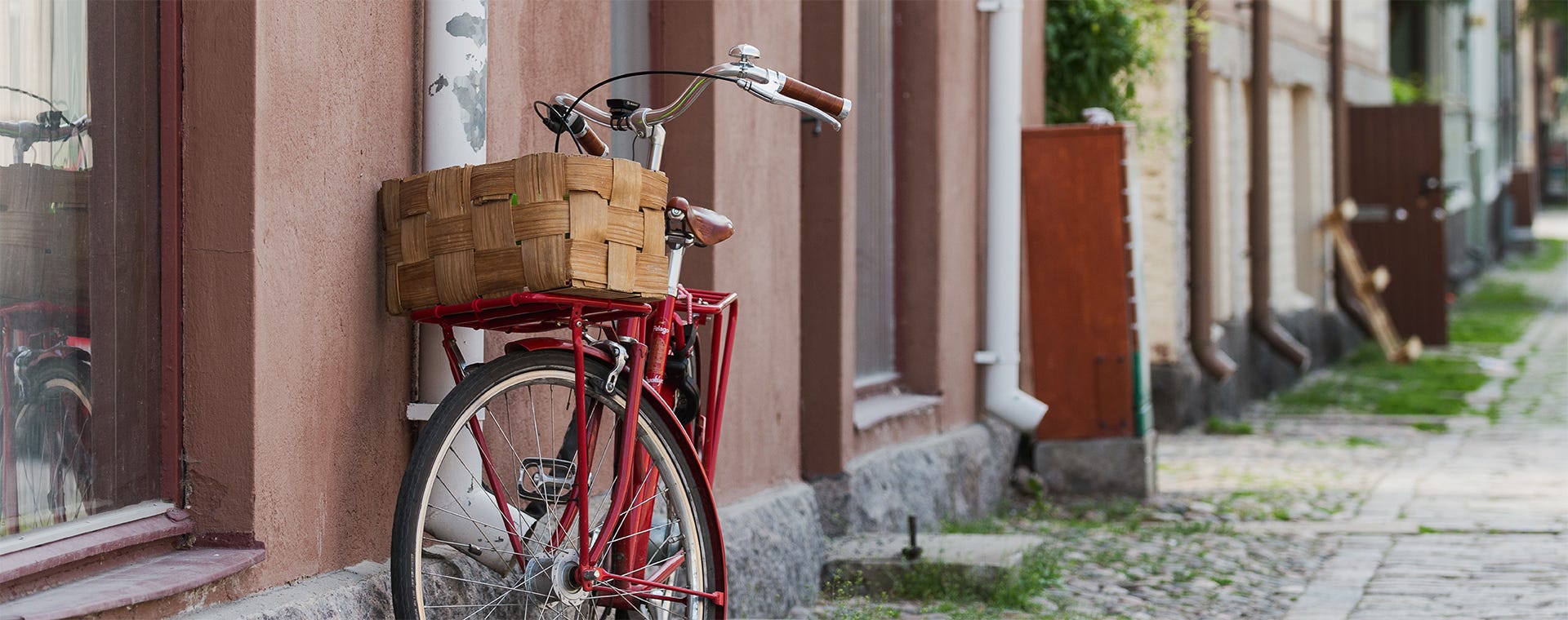 Vanhan rakennuksen seinää vasten nojaava punainen polkupyörä, jossa on etutelineellä puinen kori. Taustalla näkyy kivipäällysteinen katu ja pastellisävyisiä taloja Turun idyllisessä kaupunkimaisemassa.