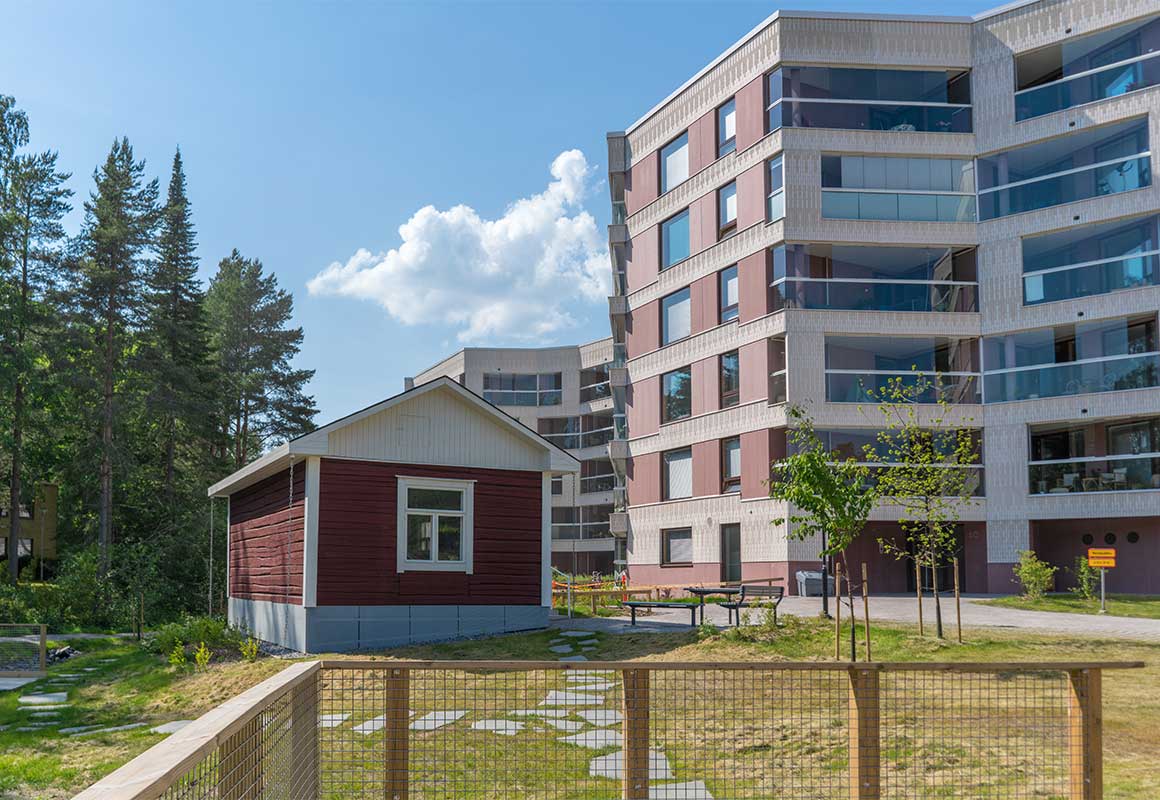 Punainen hirsimökki ja moderni kerrostalo vierekkäin vehreällä piha-alueella kesäpäivänä.