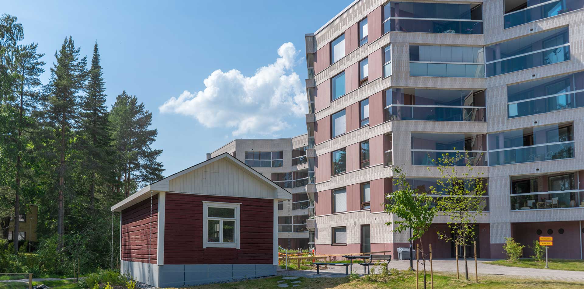 Punainen hirsimökki ja moderni kerrostalo vierekkäin vehreällä piha-alueella kesäpäivänä.