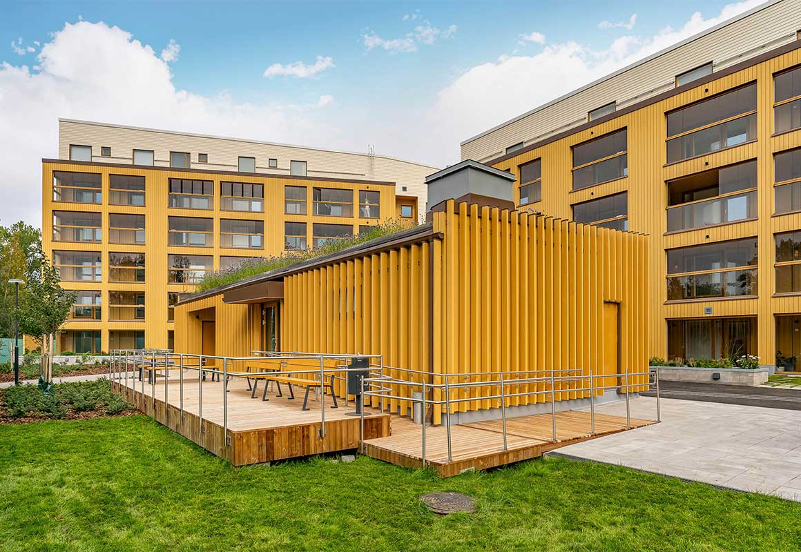 Moderni keltainen kerrostalo Espoon Tuulikello 2 -uudiskohteessa. Etualalla puurakenteinen piharakennus, jonka katolla viherrakennelma. Hyvin hoidetut viheralueet ja kulkureitit ympäröivät rakennusta.