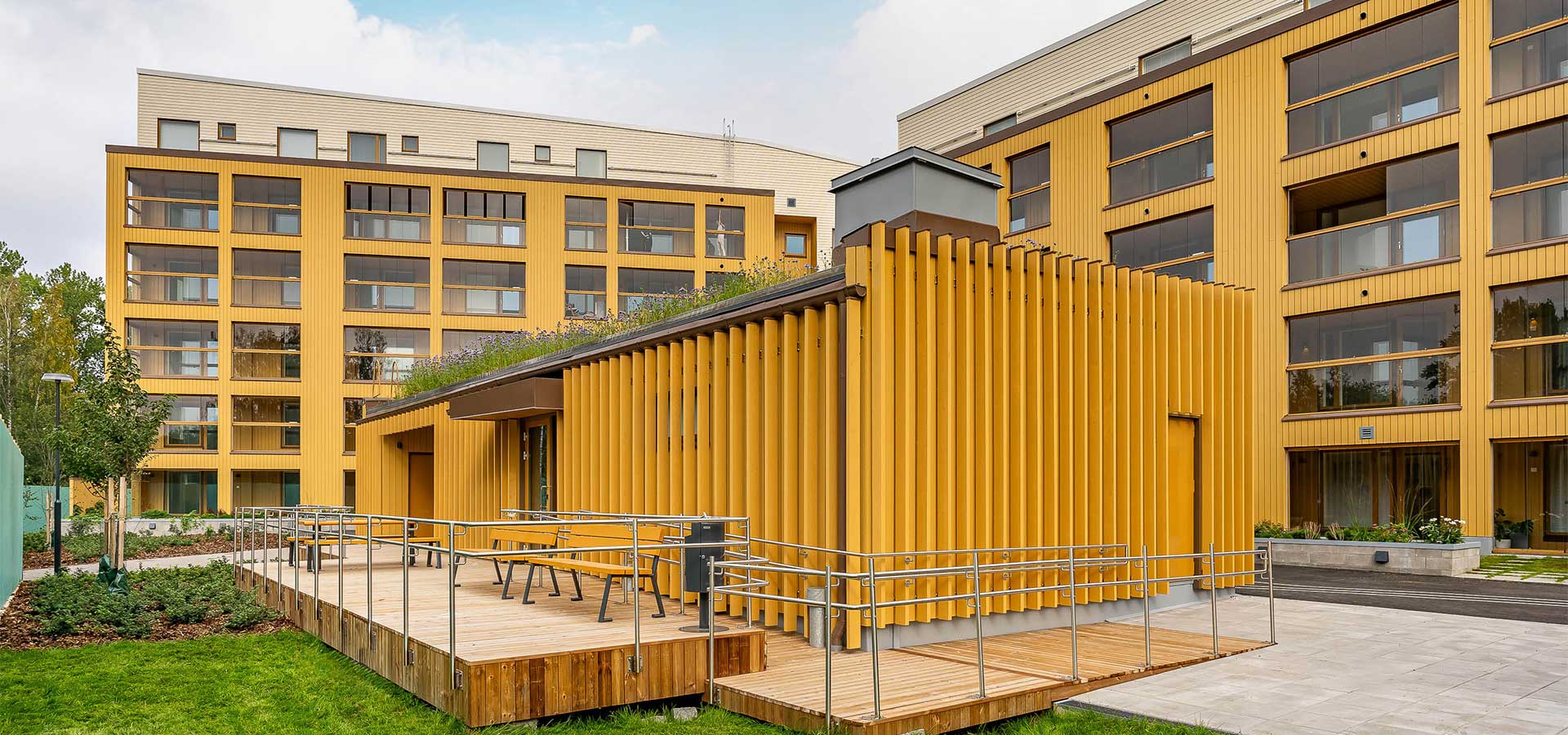 Moderni keltainen kerrostalo Espoon Tuulikello 2 -uudiskohteessa. Etualalla puurakenteinen piharakennus, jonka katolla viherrakennelma. Hyvin hoidetut viheralueet ja kulkureitit ympäröivät rakennusta.