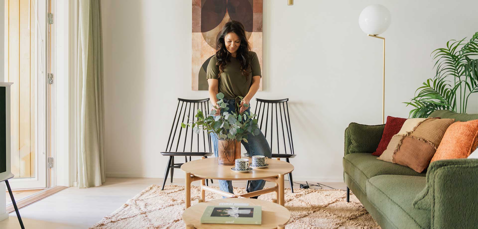 Nainen somistaa olohuonetta asettelemalla vihreitä lehtiä maljakkoon pyöreän puupöydän äärellä. Huoneessa on vihreä sohva, lämminvärinen matto ja kaksi mustaa pinnatuolia seinän vieressä.