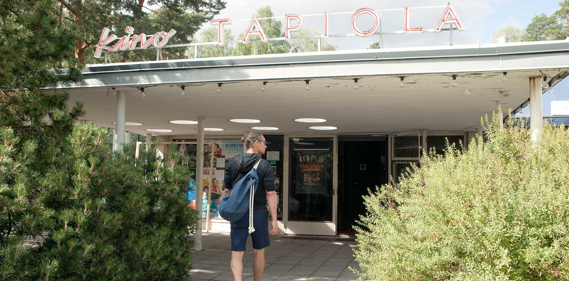 Henkilö kävelemässä kohti Kino Tapiola -elokuvateatterin sisäänkäyntiä. Rakennuksen katolla neonvalokyltti "Kino Tapiola", jota ympäröivät vehreät pensaat ja korkeat puut luovat rauhallisen ympäristön.