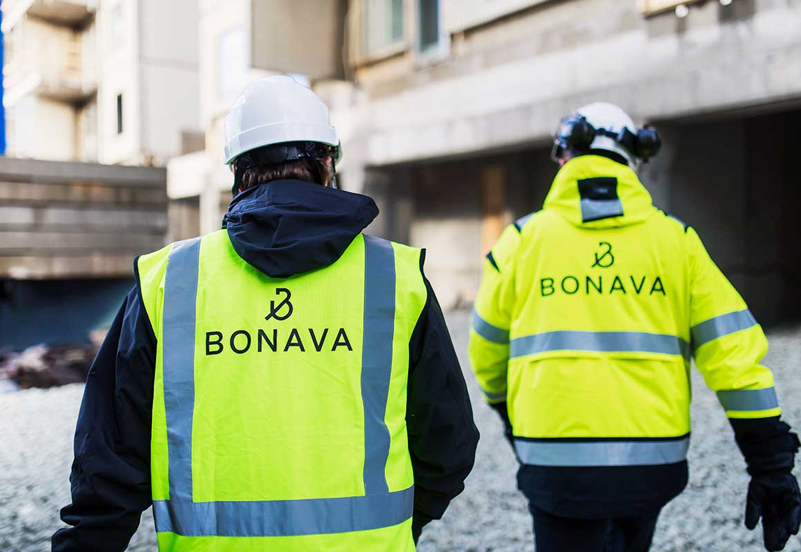 Kaksi Bonavan työntekijää kulkee rakennustyömaalla huomiovaatteissa ja suojakypärissä, taustalla rakenteilla oleva rakennus.
