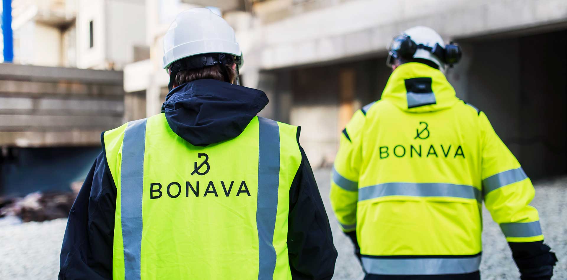 Kaksi Bonavan työntekijää kulkee rakennustyömaalla huomiovaatteissa ja suojakypärissä, taustalla rakenteilla oleva rakennus.