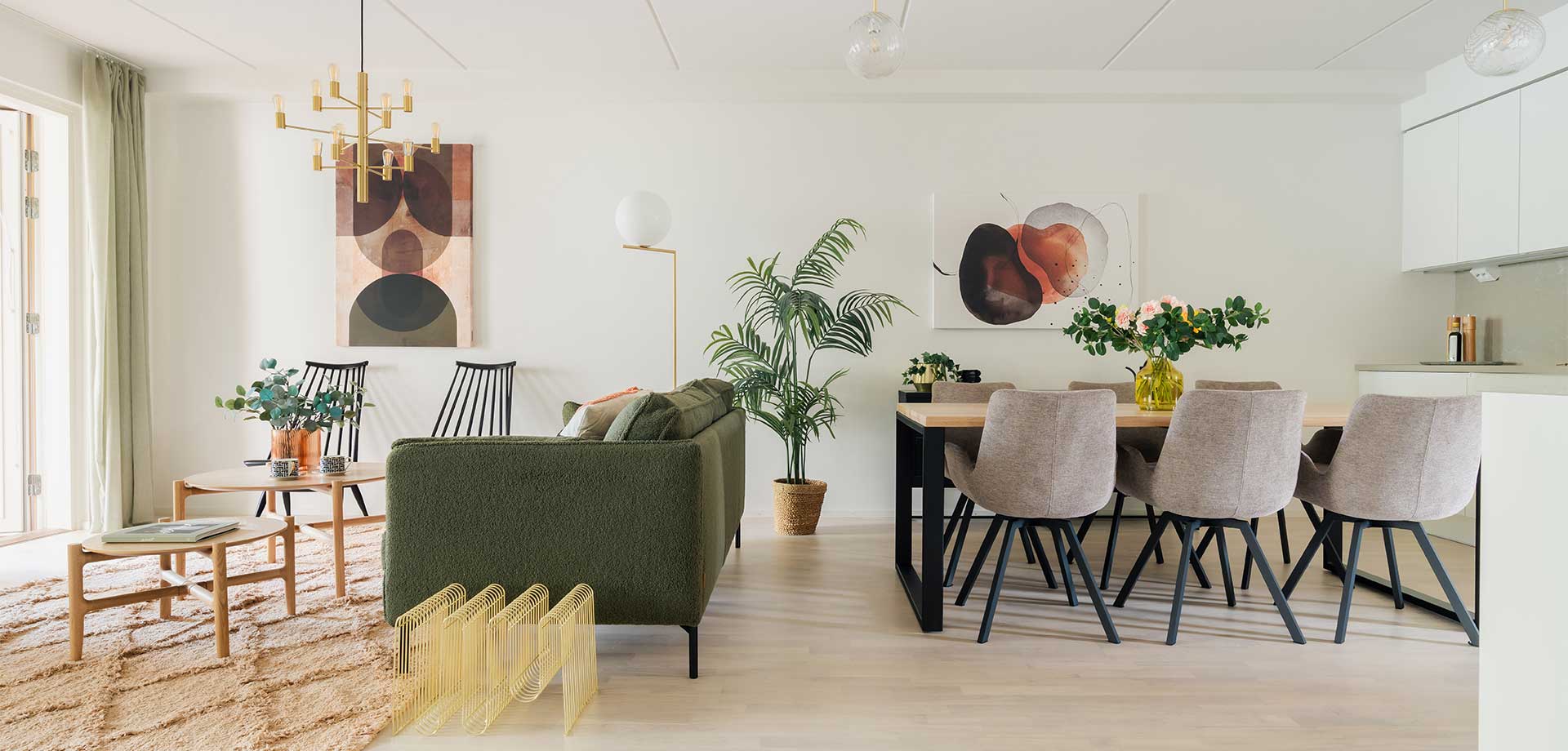 Valoisa ja moderni olohuone- ja ruokailutila, jossa on vihreä sohva, puinen ruokapöytä ja vaaleat tuolit. Koristeltu tauluilla, viherkasveilla ja näyttävällä kattovalaisimella. Avoin ja raikas sisustus, jossa on kodikas tunnelma.