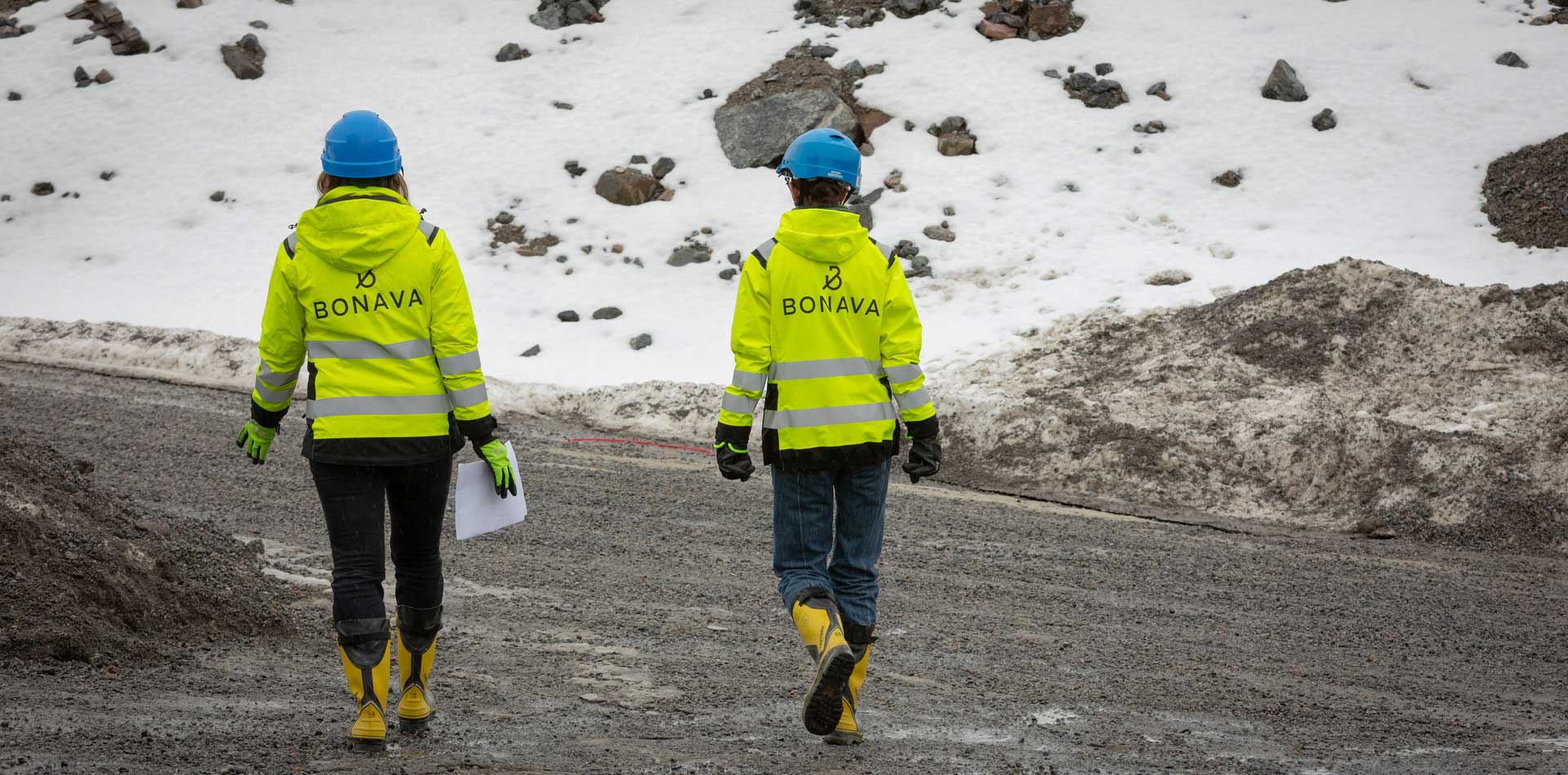 Kaksi Bonavan työvaatteisiin ja kypäriin pukeutunutta rakennusalan ammattilaista kävelee työmaalla talvisessa ympäristössä.