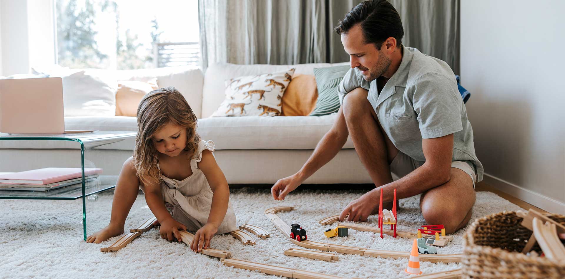 sä ja pieni tyttö rakentavat yhdessä puisia junaratoja vaalealla matolla olohuoneessa. Taustalla näkyy vaalea sohva, jossa on tyynyjä tiikeri- ja yksivärisillä kuoseilla. Rentoa ja leikkisää yhdessäoloa valoisassa kodissa.