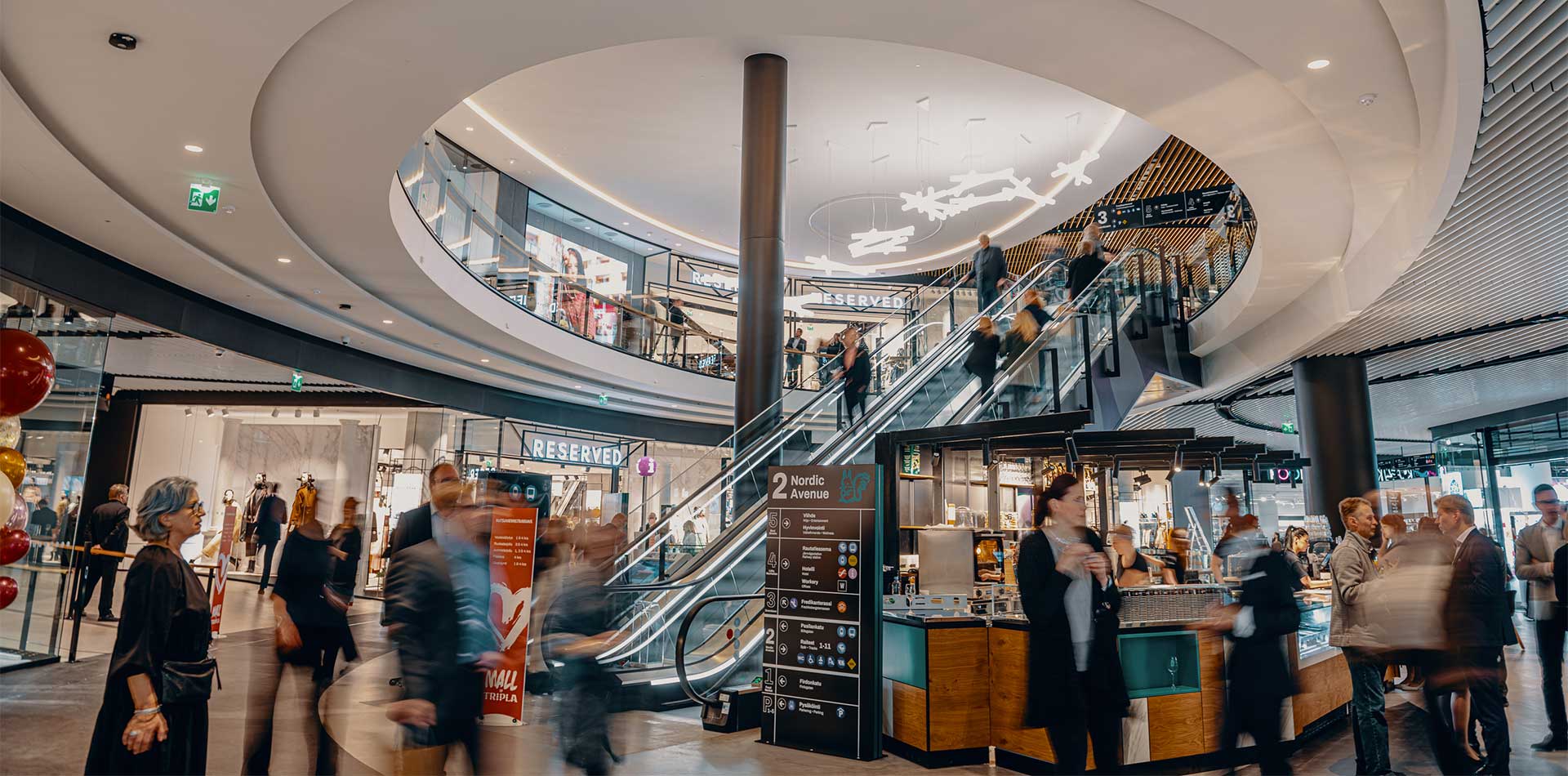 Vilkas kauppakeskus, jossa ihmiset liikkuvat liukuportaiden ja myymälöiden ympärillä useassa kerroksessa, valaistuna moderneilla kattovaloilla.
