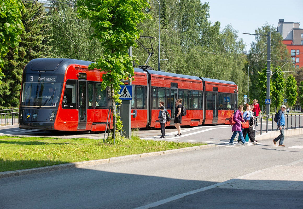 Punainen raitiovaunu pysähtyneenä Kalevanrinteen pysäkillä Tampereella, ihmisiä nousemassa kyytiin ja ylittämässä suojatietä kesäpäivänä.