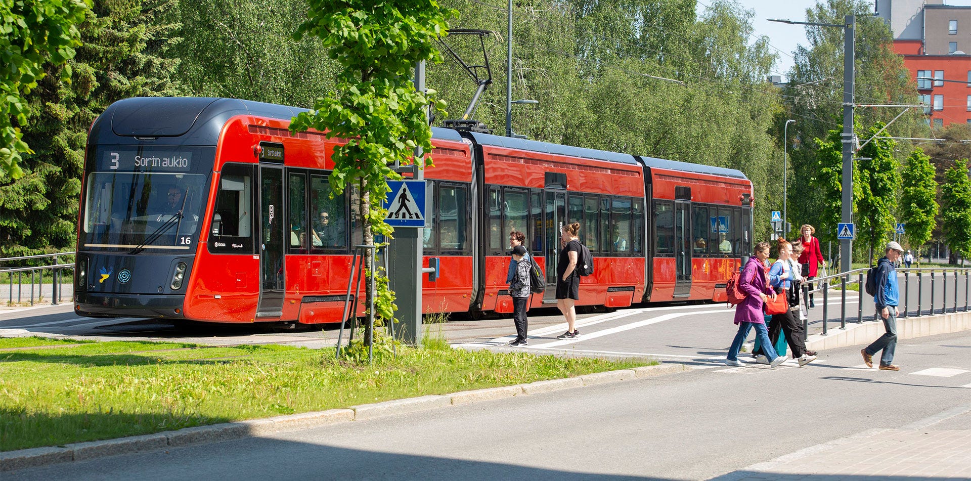 Punainen raitiovaunu pysähtyneenä Kalevanrinteen pysäkillä Tampereella, ihmisiä nousemassa kyytiin ja ylittämässä suojatietä kesäpäivänä.