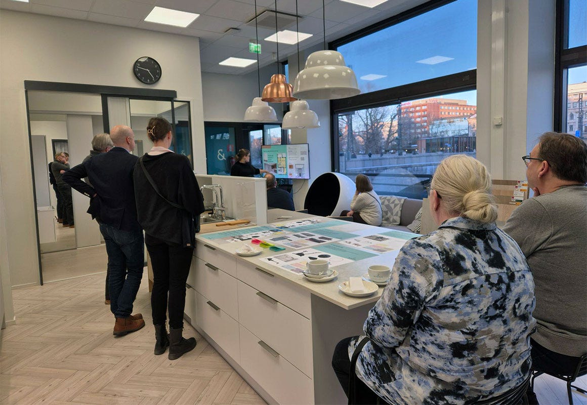 Ihmisiä Tampereen Bonavan asuntomyymälän esittelytilassa, jossa keskustellaan ja tarkastellaan sisustusvalintoja suunnittelupöydän ääressä. Pöydällä kahvikuppeja ja esitemateriaaleja.