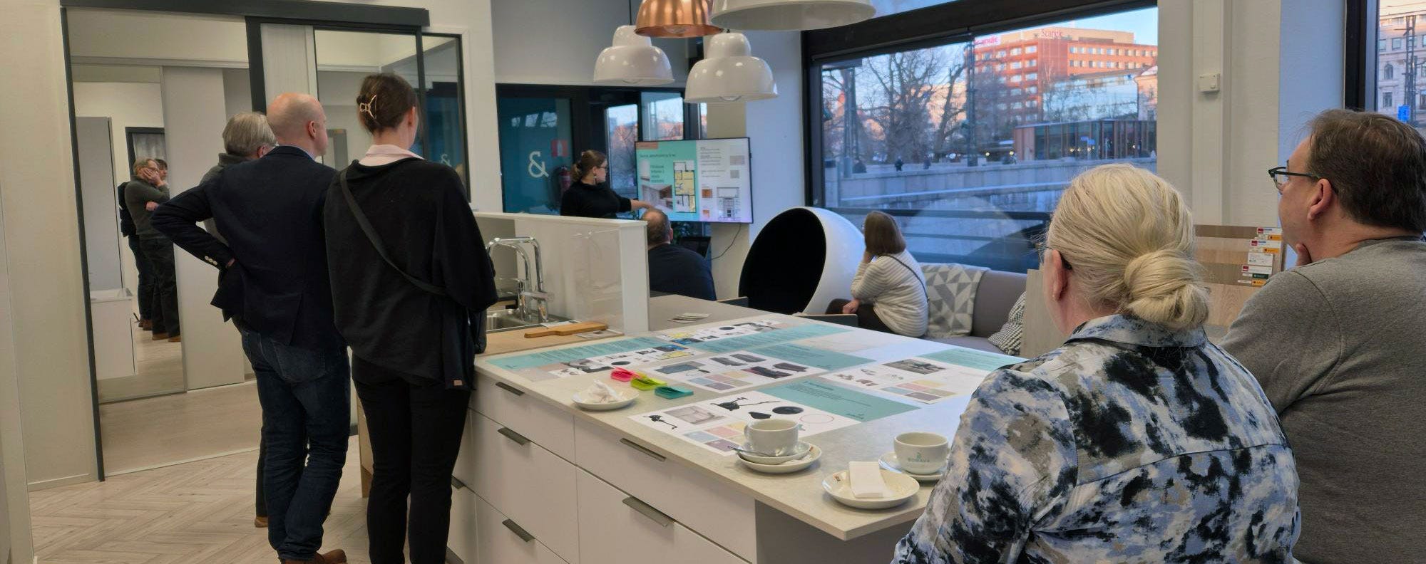 Ihmisiä Tampereen Bonavan asuntomyymälän esittelytilassa, jossa keskustellaan ja tarkastellaan sisustusvalintoja suunnittelupöydän ääressä. Pöydällä kahvikuppeja ja esitemateriaaleja.