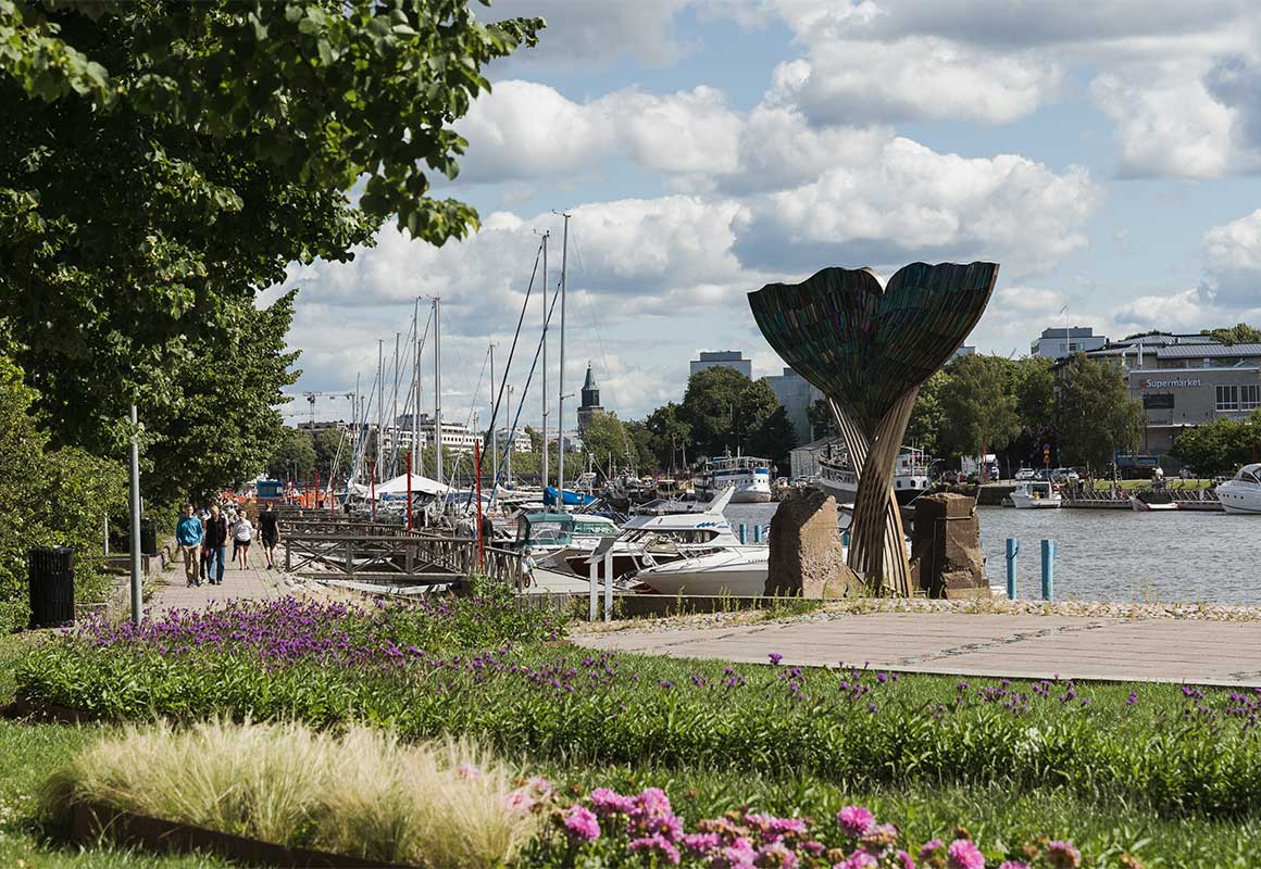 Aurajoen rantamaisema Turussa. Etualalla kävelyreitti, värikkäitä kukkia ja puistomaisuutta. Taustalla venesatama, purjeveneiden mastoja, puiden lomasta pilkottava kirkontorni ja kaupunkirakennuksia.