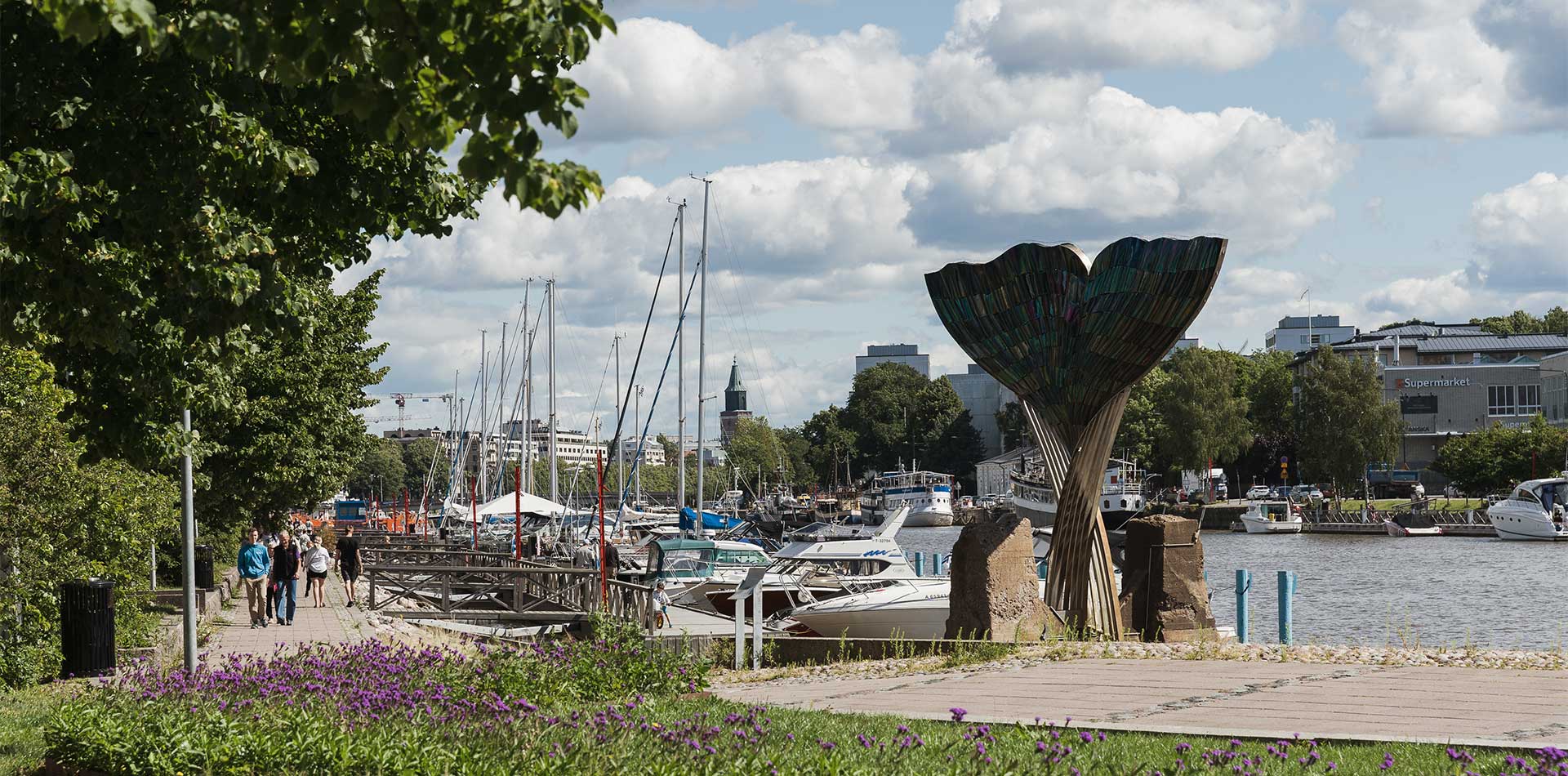 Aurajoen rantamaisema Turussa. Etualalla kävelyreitti, värikkäitä kukkia ja puistomaisuutta. Taustalla venesatama, purjeveneiden mastoja, puiden lomasta pilkottava kirkontorni ja kaupunkirakennuksia.