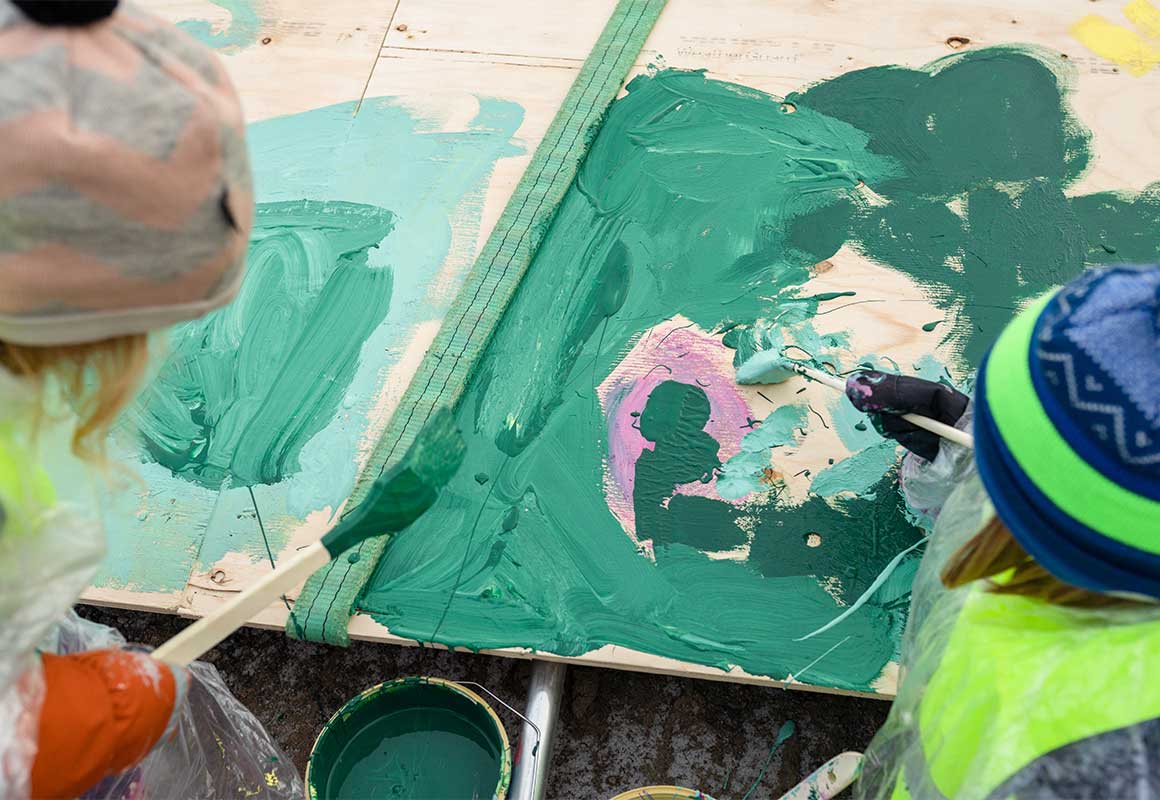 Lähikuva lasten käsistä, jotka maalaavat vihreällä ja vaaleanpunaisella maalilla rakennustyömaan puuaitaa.