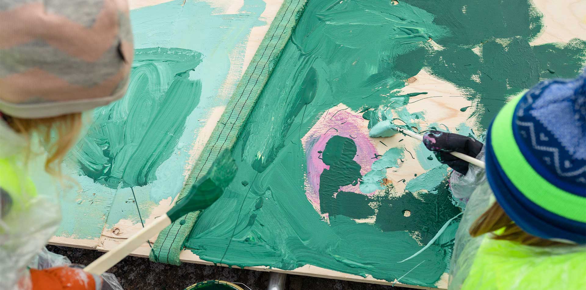 Lähikuva lasten käsistä, jotka maalaavat vihreällä ja vaaleanpunaisella maalilla rakennustyömaan puuaitaa.