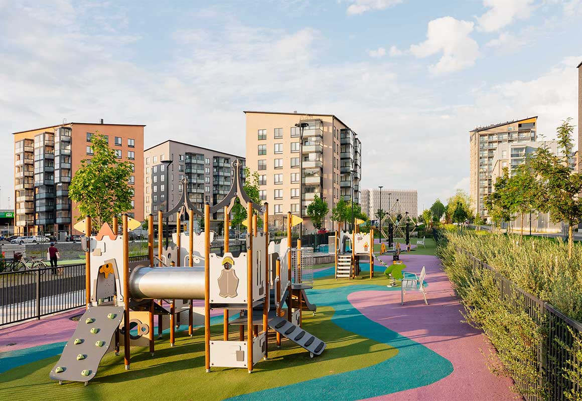 Leikkipuisto, jossa on värikäs kumipintainen leikkialue ja modernit kerrostalot taustalla aurinkoisena päivänä Turun Kirstinpuistossa.
