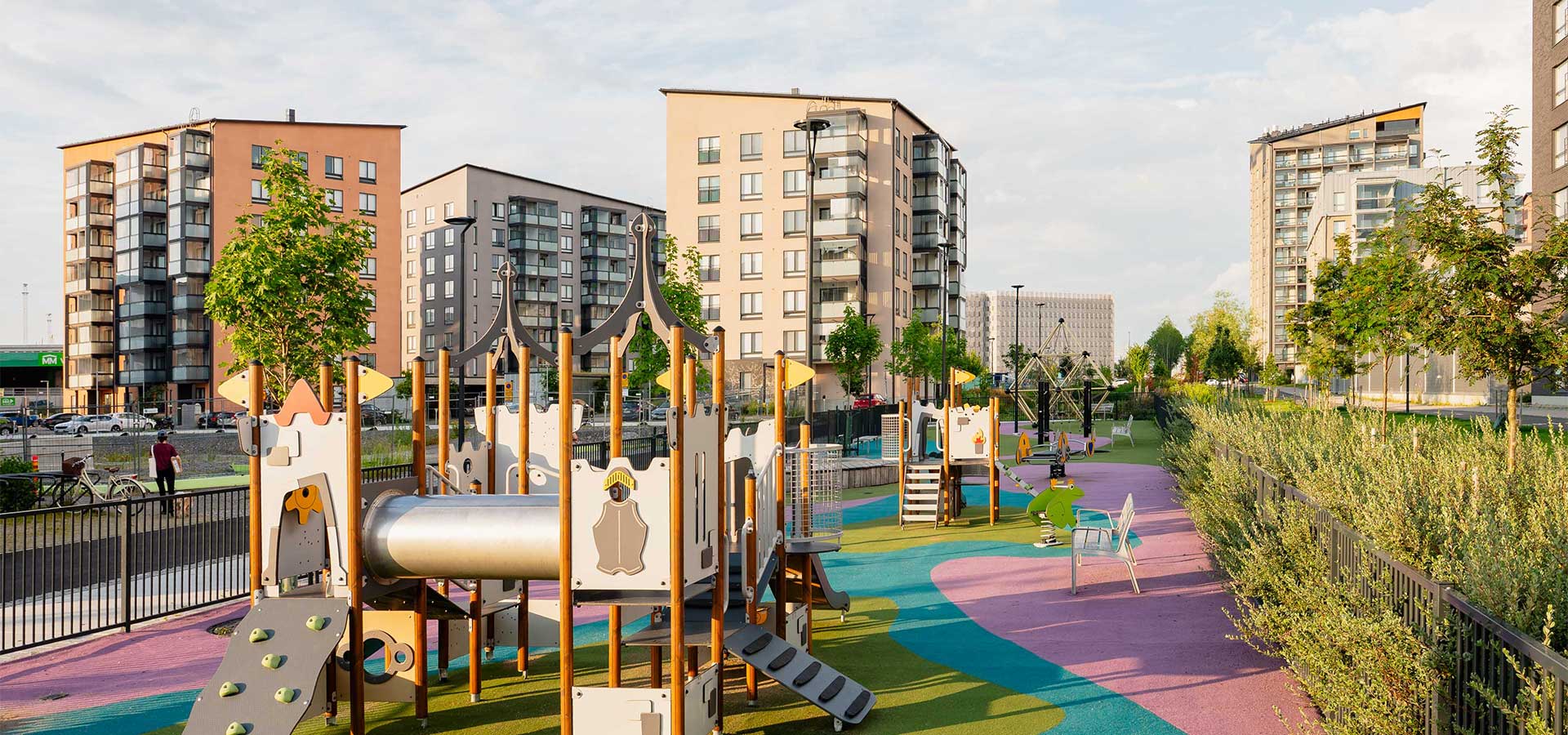 Leikkipuisto, jossa on värikäs kumipintainen leikkialue ja modernit kerrostalot taustalla aurinkoisena päivänä Turun Kirstinpuistossa.