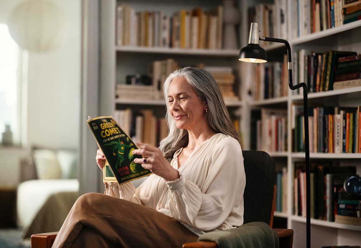 Nainen istuu nojatuolissa ja lukee lehteä kodikkaassa olohuoneessa kirjahyllyn äärellä.