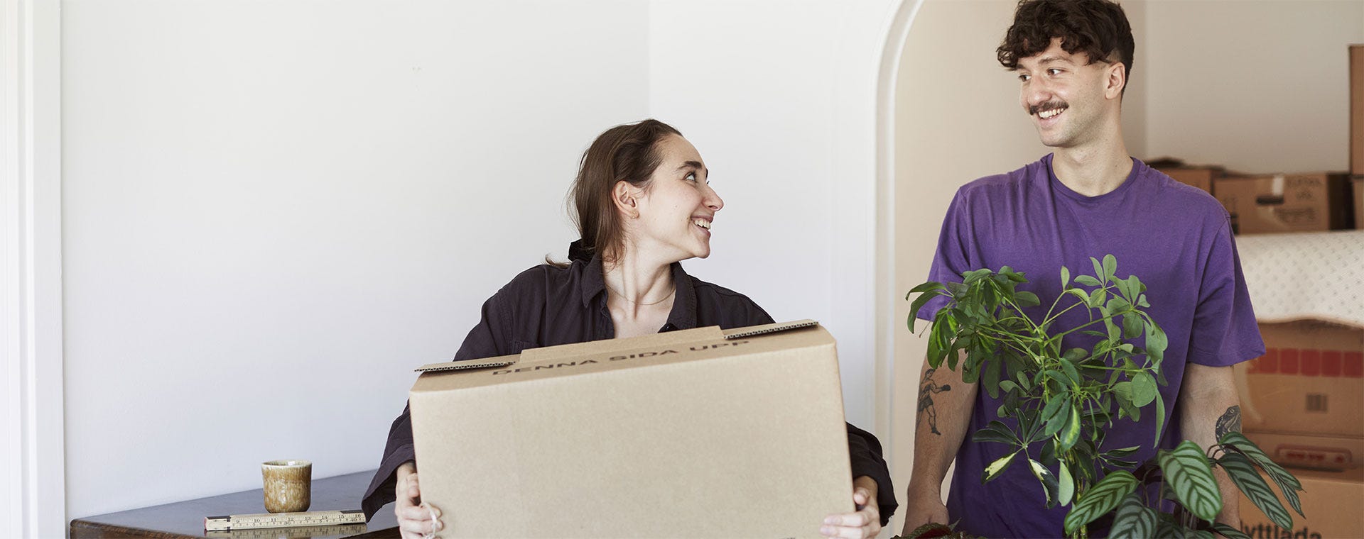 Nuori pariskunta muuttaa uuteen kotiin iloisin mielin. Toinen kantaa muuttolaatikkoa ja toinen viherkasvia, taustalla näkyy laatikoita ja huonekaluja. Kuvassa välittyy uuden kodin innostus ja yhteinen elämänvaihe.