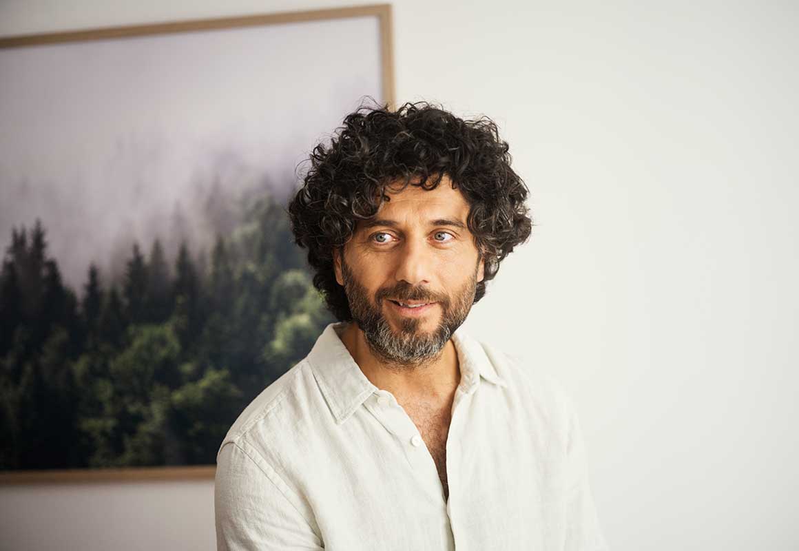 Mies seisoo sisätilassa ja katsoo kameraan, taustalla seinällä metsäaiheinen taulu.